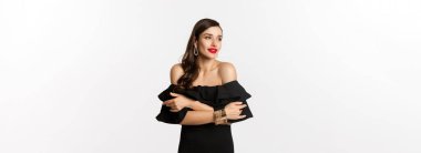 Güzellik ve moda konsepti. Siyah elbiseli, makyajlı zarif ve güzel bir kadın. Kendini kucaklıyor ve bakışlarını başka tarafa çeviriyor. Beyaz arka planda duruyor..