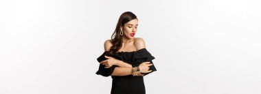 Güzellik ve moda konsepti. Siyah elbiseli, kırmızı dudaklı, şehvetli ve çekici bir kadın, kendini kucaklıyor ve nazikçe aşağıya bakıyor, beyaz arka planda duruyor..