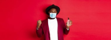 Covid-19, karantina ve tatil konsepti. Neşeli siyah adam kazanmaktan, hedefe ulaşmaktan, antiseptik göstermekten, tıbbi maske takmaktan, kırmızı arka planda durmaktan memnun..
