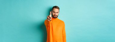 Ciddi sakallı adam kaşlarını çatıyor, hareket yasağı koymak için parmağını sallıyor, bir şeyi yasaklıyor, hayal kırıklığını dile getiriyor, açık mavi arka planda seninle aynı fikirde değil..