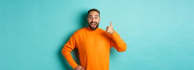 İyi iş çıkardın. Yakışıklı sakallı adam baş parmağını gösterip, iyi işi övüyor, mükemmel bir ürün öneriyor, onaylıyor ve onaylıyor, gülümsüyor, açık mavi arka planda duruyor..