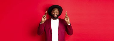 Umut dolu Afrikalı Amerikalı bir adam dilek tutuyor, şans diliyor ve iyimser gülümsüyor, kırmızı parti geçmişine karşı duruyor..