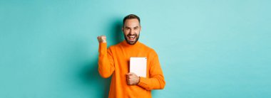 Heyecanlı yetişkin adam dizüstü bilgisayarını tutuyor ve yumruk pompası yapıyor, galibiyetin sevinci, turkuaz arka planda tatmin olmuş duruyor..