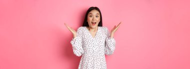 Mutlu ve şaşırmış Asyalı kadın seviniyor, ellerini açıyor ve hayretle nefes nefese, heyecan ve inançsızlıkla bakıyor, pembe arka planda duruyor..