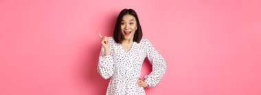Heyecanlı güzel Asyalı kız sağ üst köşeyi işaret ediyor, şaşırmış ve kameraya bakıyor, mağaza tanıtım teklifini tartışıyor, pembe arka planda duruyor..