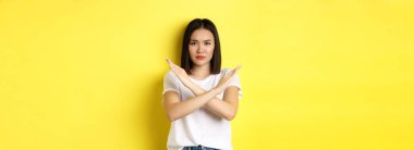 Ciddi ve iddialı Koreli kadın dur işareti yapıyor, kaşlarını çatıyor ve hayır diyor, eylemi yasaklıyor, kötü bir şeyi onaylamıyor, sarı arka planda duruyor..