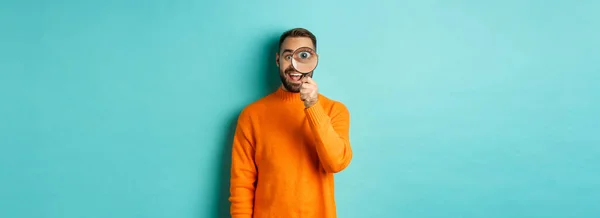 Cheerful man searching for something, looking through magnifying glass and smiling happy, standing over blue background.
