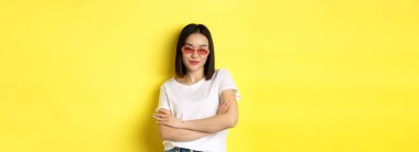 Moda ve yaşam tarzı konsepti. Şımarık ve kendinden emin güneş gözlüklü Asyalı kadın kameraya kendinden emin bakıyor, sarı arka planda duruyor..