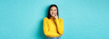 Güzel görünümlü, şık giyinmiş, dudağına dokunan ve mutlu yüzüyle kameraya gülümseyen, mavi arka planda duran Asyalı bir kadın resmi..