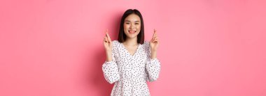 Tatlı Asyalı bir kadın dilek diliyor. Mutlu bir şekilde gülümsüyor ve şans diliyor. Pembe arka planda bekliyor..