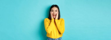 Neşeli ve şaşırmış Asyalı kadın terfiyi kontrol ediyor, hayrete düşmüş, mavi arka planda duruyor..