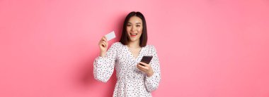 Çekici genç Asyalı kadın online sipariş veriyor, kredi kartı ve cep telefonu tutuyor, internet alışverişi yapıyor, pembe arka planda mutlu duruyor..