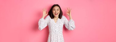Hoş, genç Asyalı bir kadın iyi bir işaret veriyor, övgüler yağdırıyor ve onaylıyor, tatmin olmuş görünüyor, pembe arka plana karşı duruyor..