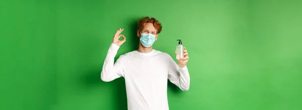 Covid-19, virus and social distancing concept. Smiling young man with red hair, wearing face mask from coronavirus, showing okay sign and hand sanitizer, green background.
