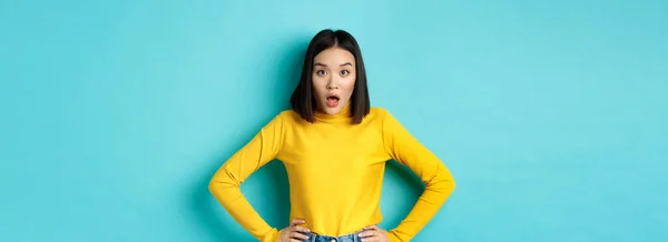 Portrait of surprised asian woman drop jaw, listening to big news, looking amazed at camera, standing in yellow sweater against blue background.