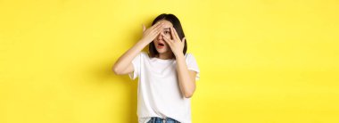 Şaşırmış Asyalı kız şaşırmayı bekliyor, parmaklarının arasından bakıyor, mutlu bir şekilde gülümsüyor, sarı arka planda duruyor..