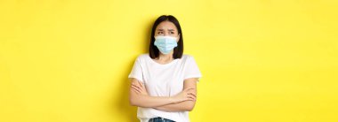 Covid, sağlık ve salgın hastalık konsepti. Beyaz tişörtlü ve tıbbi maskeli Asyalı bir kadın göğsünde çapraz kollar ve dalgın ve endişeli bir şekilde yukarı bakıyor..