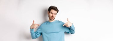 Sakallı genç adam kendine güveniyor, kendini gösteriyor ve kendini beğenmiş bir ifade takınıyor, beyaz arka planda havalı ve küstah duruyor..