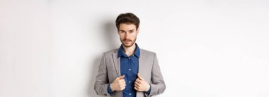 Sakal düzeltme kıyafeti giymiş, kameraya küstah bakışlı, hazır ve kararlı, beyaz arka planda duran kendine güvenen genç bir adam..