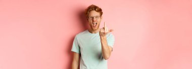 Arrogant and rude redhead man in glasses dont give a fuck, showing middle fingers at camera and frowning, standing over pink background.