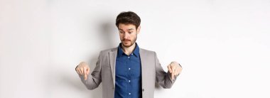 Şaşkın iş adamı yukarı kaş kaldırarak aşağı bakıyor ve tuhaf bir şey görüyor, takım elbiseli beyaz arka planda duruyor..