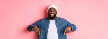 Yakışıklı afro-amerikan adam parmaklarını eğip, gülümsüyor ve tanıtım teklifini sunuyor, pembe arka planda duruyor..