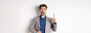 Heyecanlı takım elbiseli işadamı iyi fikir sunuyor, Eureka işaretinde parmağını kaldırıyor ve komik görünüyor, öneriler söylüyor, beyaz arka planda duruyor..