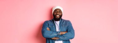 Yakışıklı hip hop stili bereli, kot gömlekli, kendine güvenen, göğsünde çapraz kollar olan ve pembe arka planda kameraya bakan siyah bir adam..