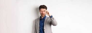 İş adamı küçük bir şey görmeye çalışıyor, parmaklarıyla küçük bir şey gösteriyor, beyaz arka planda takım elbise içinde duruyor..