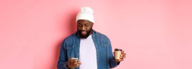 Tarz sahibi siyahi bir hippi kahve içiyor, telefonla mesaj okuyor, pembe arka planda dikiliyor..