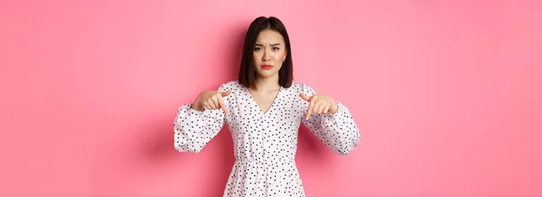 Disappointed asian female model pointing fingers down at promo offer, frowning and staring at camera upset, standing over pink background.