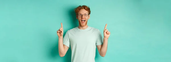 Surprised young man with ginger hair, wearing glasses and t-shirt, gasping in awe and pointing fingers up at promo deal, standing over mint background.