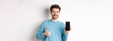 Gülümseyen genç adam boş akıllı telefon ekranı gösteriyor ve başparmak yukarı, iyi uygulama övgü, mobil uygulama öneriyor, beyaz arkaplan üzerinde duran.