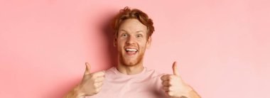 Kızıl saçlı ve sakallı neşeli bir adama yaklaş, başparmağını kaldır ve gülümse, evet de, pembe arka planda duran havalı bir şeyi onayla ve öv..