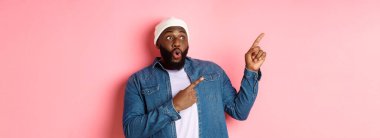 Şapkalı ve kot gömlekli etkileyici hippi siyahi adam sol üst köşedeki logoya bakıyor ve işaret ediyor, şaşkın şaşkın bakıyor, pembe arka planda duruyor..