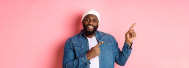 Mutlu Afro-Amerikalı adam sol üst köşeyi işaret ediyor, tanıtım logosu gösteriyor, gülümsüyor memnun, kot ceketli bere giyiyor, pembe arka planı var..