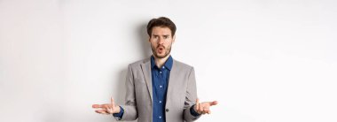 Kafası karışmış ve şok olmuş işadamı elini kaldırıyor ve ne olduğunu soruyor, kaşlarını çatmış, açıklama bekliyor, beyaz arka planda takım elbise içinde duruyor..