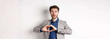 Yakışıklı bir adam aptal görünüyor, kalbi gösteriyor imzalıyor ve dudaklarını büzüyor, sevgilisinden öpücük bekliyor, beyaz arka planda takım elbise içinde duruyor..