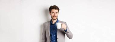 Takım elbiseli yakışıklı işadamı kahve ya da ofis kupasından çay içiyor, beyaz arka planda duruyor..