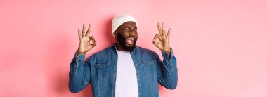Hipster bereli tatmin olmuş Afro-Amerikan bir adam, onaylama işaretleri gösteriyor, iyi bir teklifi övüyor ve evet diyor, mutlu gülümsüyor, pembe arka planda duruyor..