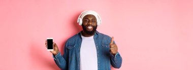 Mutlu hipster adam kulaklıkla müzik dinliyor ve mobil ekranda gülümsüyor, tatmin olmuş bir şekilde gülümsüyor, müzik listesi gibi, pembe arka planda duruyor..