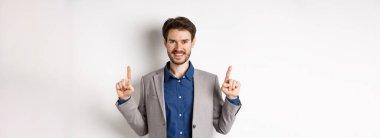 Başarılı iş adamı parmaklarını havaya kaldırıyor, gülümsüyor ve şirket logosunu gösteriyor, beyaz arka planda takım elbise içinde duruyor..