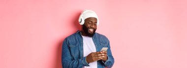Yakışıklı, şık siyahi adam kulaklıkla müzik dinliyor ve akıllı telefondan mesaj yazıyor, mutlu gülümsüyor, pembe arka planda duruyor..