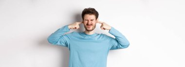 Sinirli genç adam korkunç seslerden kulaklarını tıkar, rahatsız edici gürültüden rahatsız olur, hoşnutsuz olur, beyaz arka planda durur..