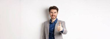 Takım elbiseli yakışıklı iş adamının resmi. Baş parmağını kaldırıyor ve gülümsüyor. Beyaz arka planda duruyor..