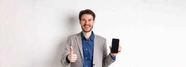 E-ticaret ve online alışveriş konsepti. Takım elbiseli tatmin olmuş iş adamı boş akıllı telefon ekranı ve baş parmak yukarı gösteriyor. İyi bir anlaşma, beyaz arka plan..