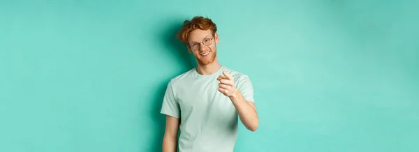 Handsome young man with ginger hair, wearing glasses and t-shirt, pointing finger at camera and smiling, choosing you, making invitation, standing over turquoise background.