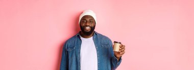 Yakışıklı, modern siyahi adam paket kahve içiyor, gülümsüyor ve kameraya tatmin olmuş görünüyor, pembe arka planda duruyor..