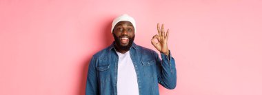 Mutlu gülümseyen siyah adam iyi işaret veriyor, onaylıyor ve övüyor. Pembe arka planda duruyor..