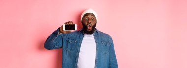 Çevrimiçi alışveriş ve teknoloji kavramı. Şaşırmış siyah adam telefon ekranını yatay olarak gösteriyor ve kameraya bakıyor heyecanlı, pembe arka planda duruyor..